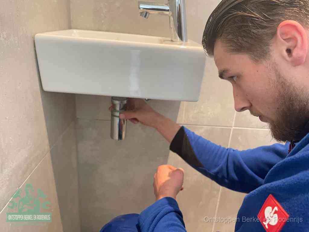 Een monteur draait de sifon onder een fonteinwasbak los om de wasbak te ontstoppen in Berkel en Rodenrijs